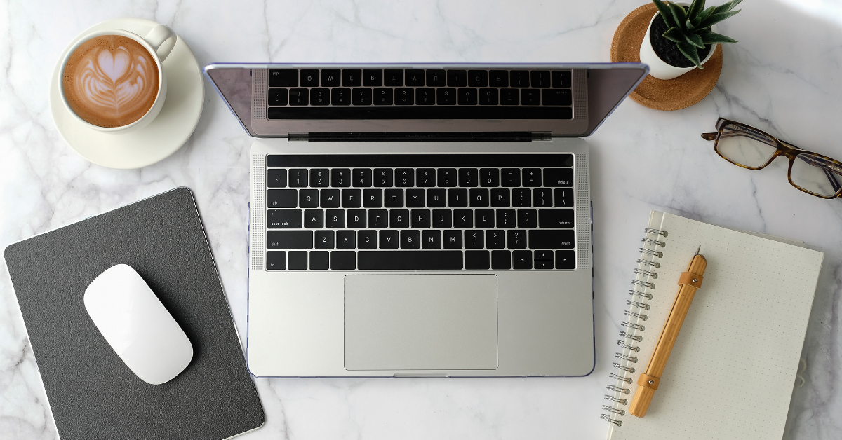 Laptop on a marble surface with a cup of coffee, mouse, notebook, and glasses.